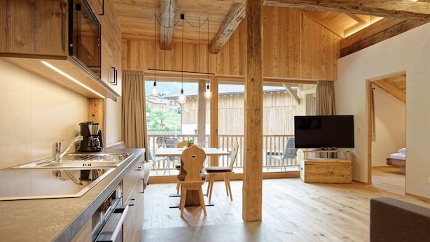 Cocina moderna con horno empotrado, cafetera y pileta. Hay un comedor con mesa y sillas, y un televisor sobre un atril de madera. El ambiente tiene pisos de madera y un balcón con vista al exterior.