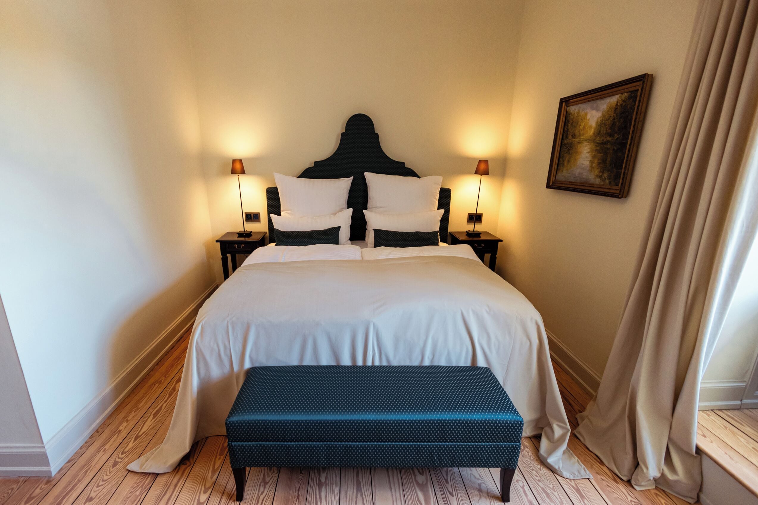 Un dormitorio ordenado con una cama grande, dos mesitas de noche con lámparas, un banco y un cuadro en la pared.