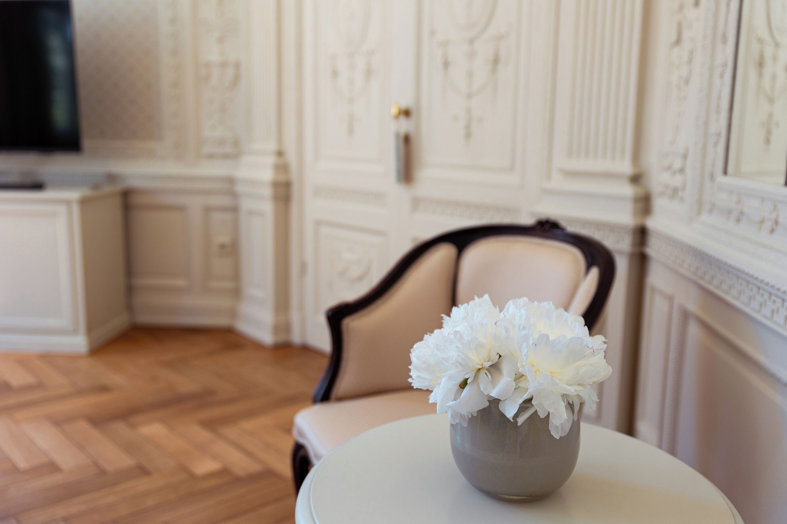 A room with a round table, a beige chair, and a vase with white flowers.