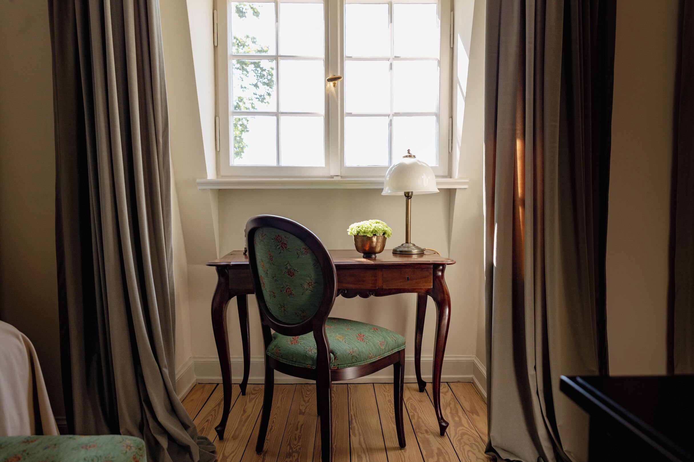 Une pièce avec un plancher en bois, un bureau avec une lampe, une chaise et une fenêtre avec des rideaux.