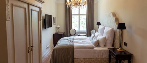A bedroom with a bed, bedside tables, a chandelier, a window with curtains, and a television on the wall.