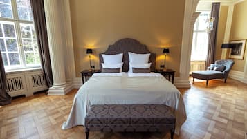 A large bed with a headboard, two bedside tables with lamps, a bench, and a chair in a room with wooden flooring and large windows.