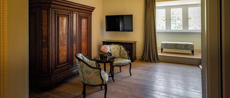Una habitación con piso de madera, un armario grande, dos sillones con estampado floral, una mesita redonda con un florero, un televisor de pantalla plana y un banco junto a una ventana con cortinas.