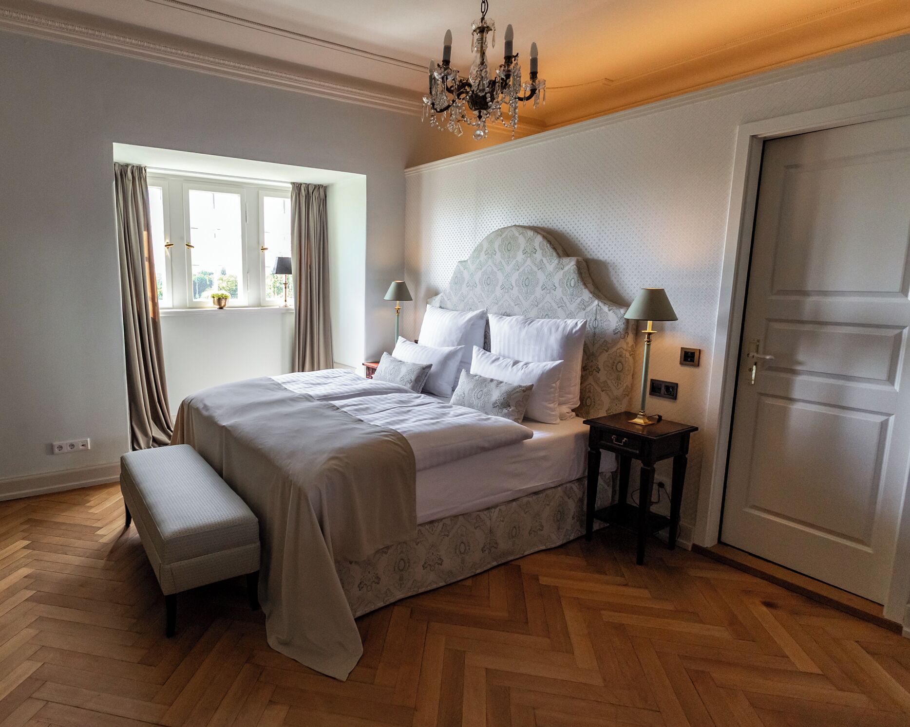 Un dormitorio con una cama grande, un banco, una mesita de noche, una araña y una puerta.