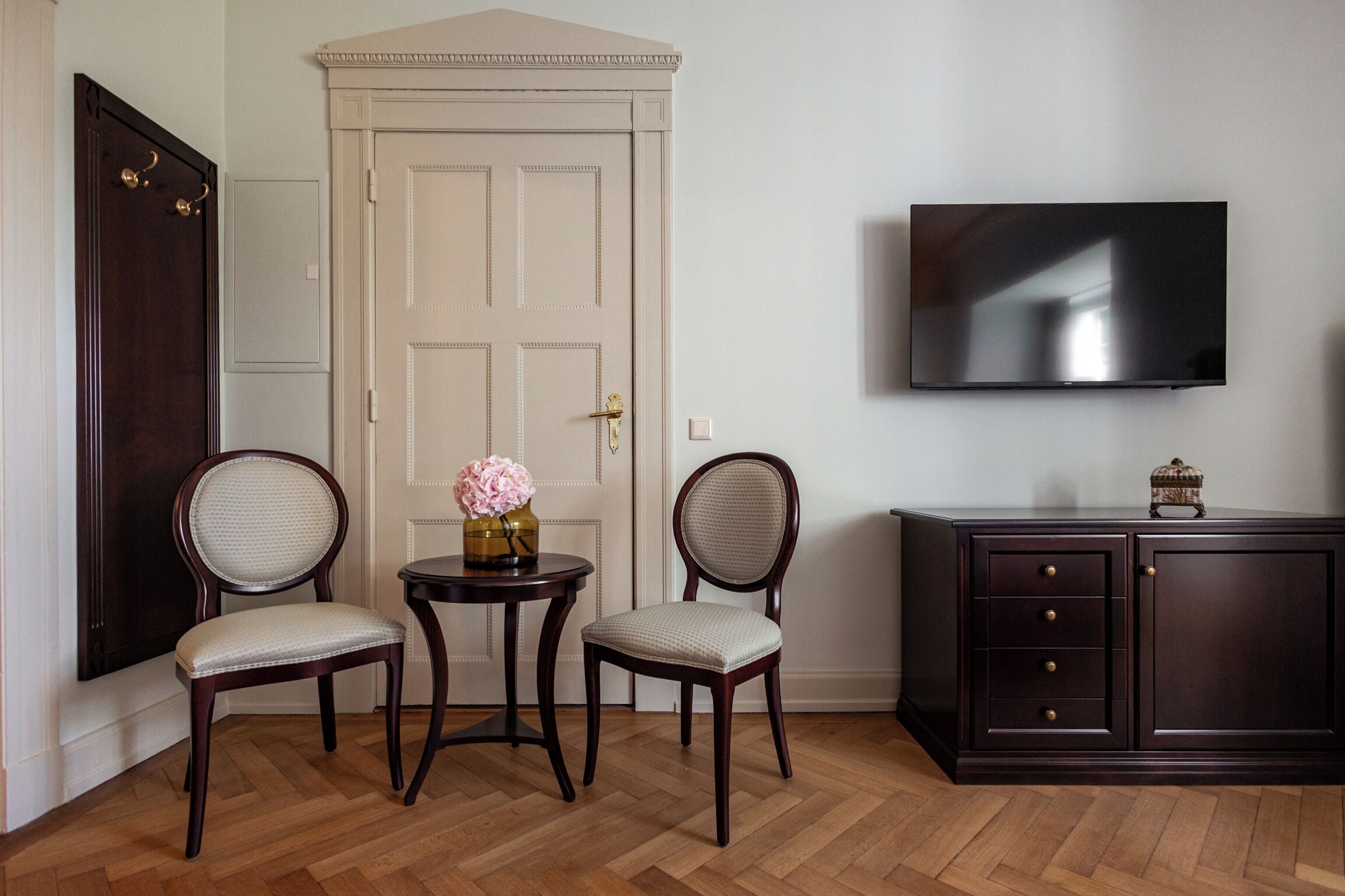 Una habitación con un mueble de madera oscura, un televisor montado en la pared y dos sillas con una mesita entre ellas.