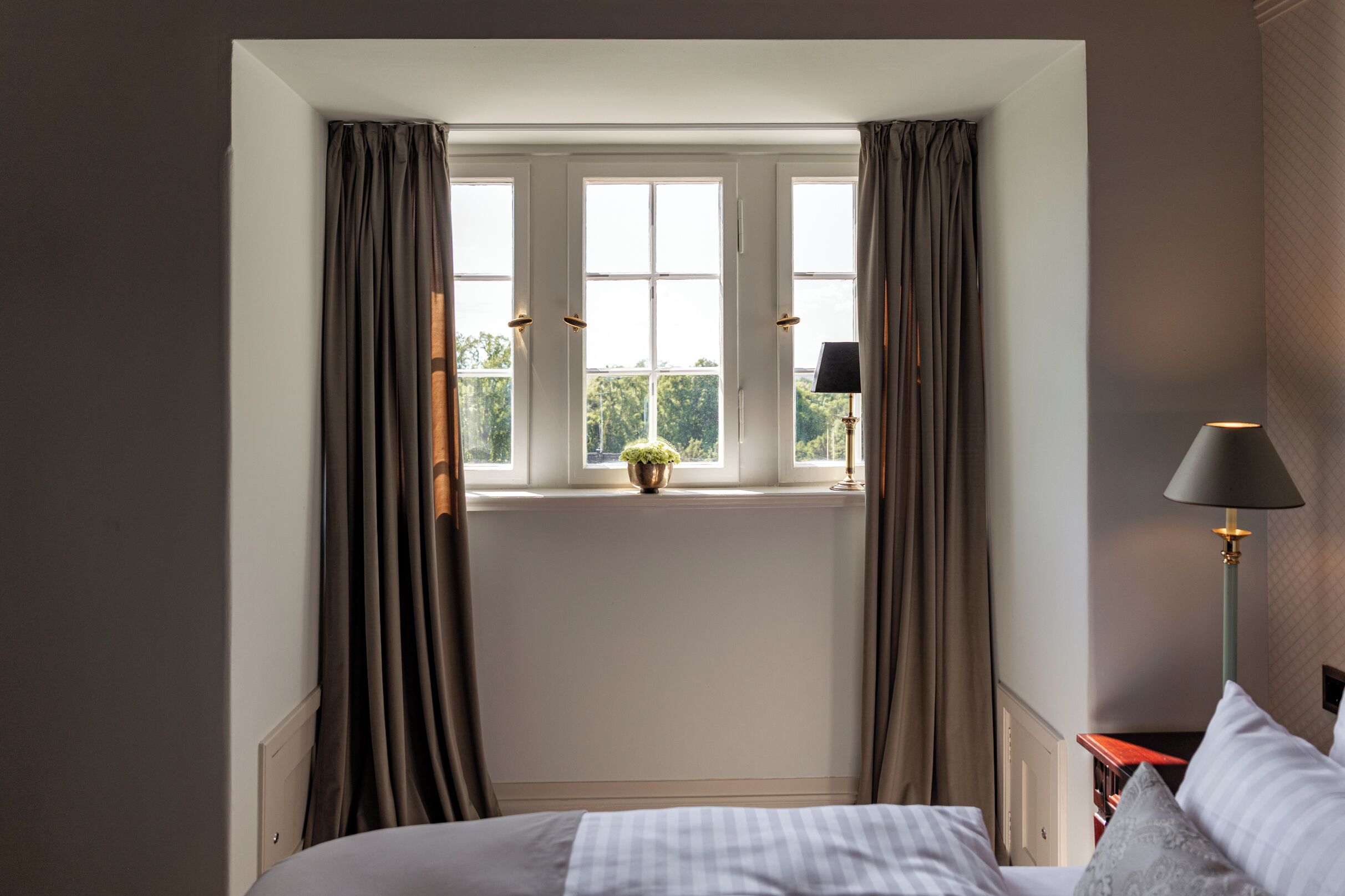 A bedroom with a large window, curtains, a bedside table with a lamp, and a bed with pillows.