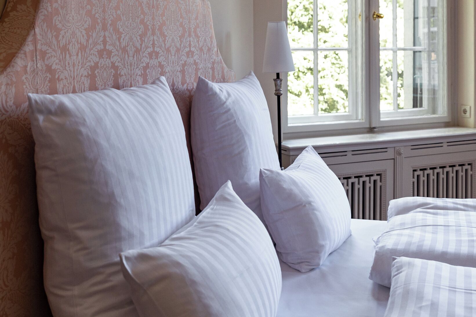 A neatly made bed with white and striped bedding, a patterned headboard, and a window with a view of greenery.