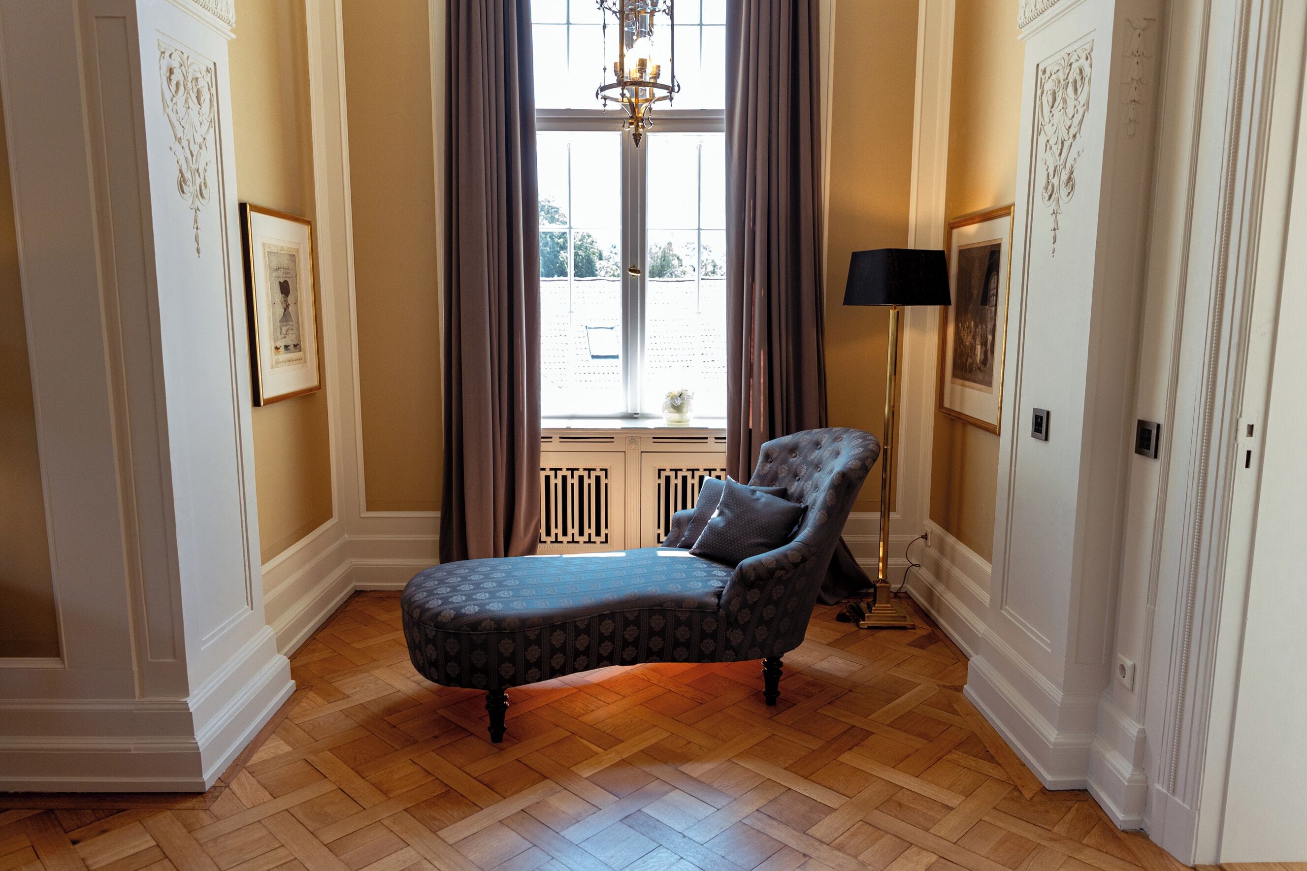 A room with a patterned ottoman, a tufted armchair, a floor lamp, and a large window with curtains.