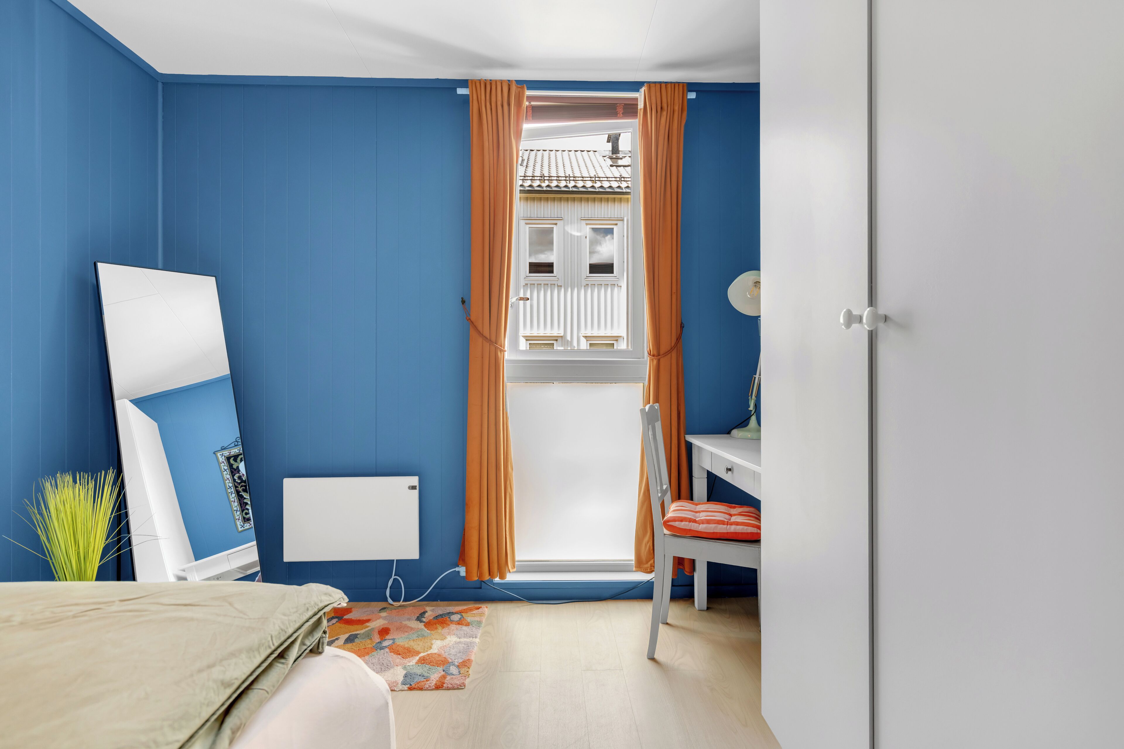 A room with blue walls, a white door, a bed, a desk with a chair, and a window with curtains.