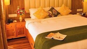 Habitación de hotel con cabecera de madera, cama con varias almohadas, mesita de noche con lámpara y un florero con flores.