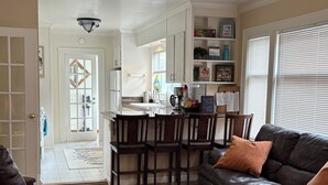 Una sala de estar con un sofá de cuero oscuro, una mesa de centro de madera y un tapete estampado. En el fondo, hay una cocina con armarios blancos y una mesa de comedor con sillas.