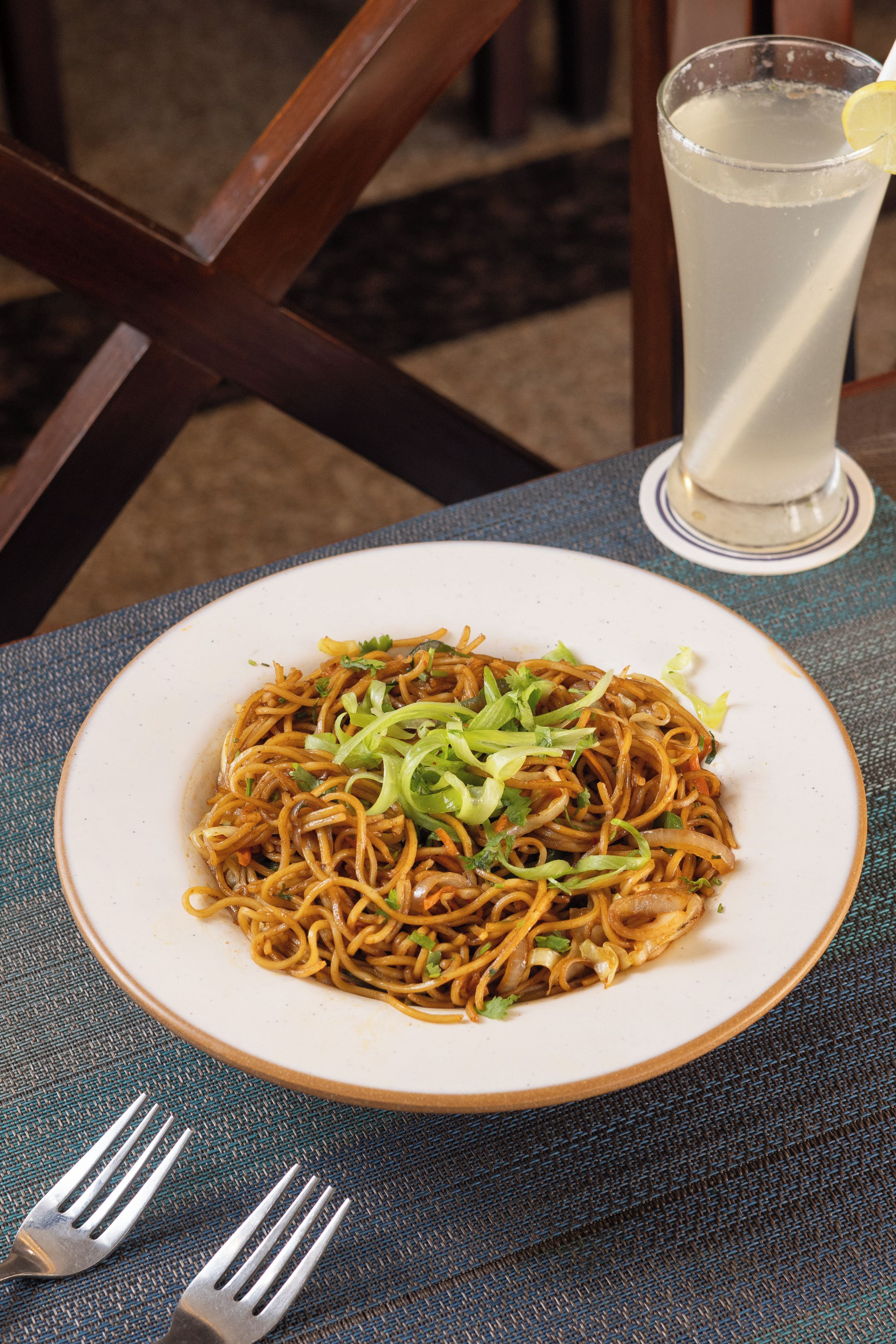 Un plato de fideos con verduras sobre una mesa, junto a un tenedor y un vaso de bebida.