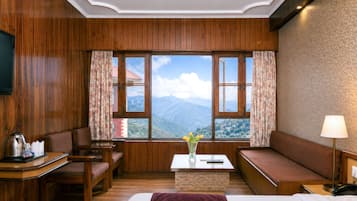 Habitación de hotel con paneles de madera, una cama, un escritorio, una silla, un sofá, una mesa con un jarrón de flores y vistas a las montañas.