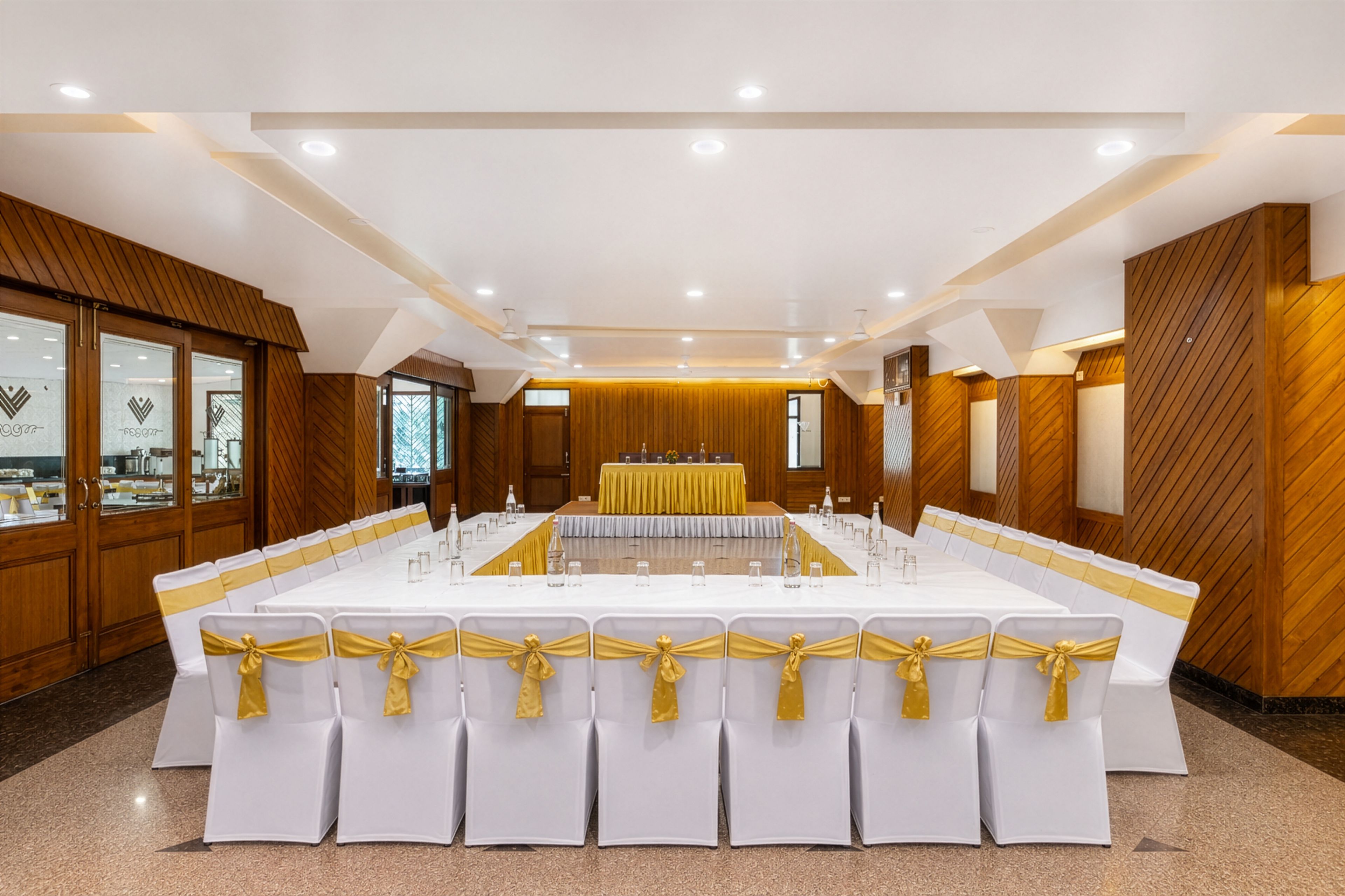Uma sala de conferências com uma mesa comprida, cadeiras brancas com fitas douradas, painéis de madeira e um teto com iluminação embutida.