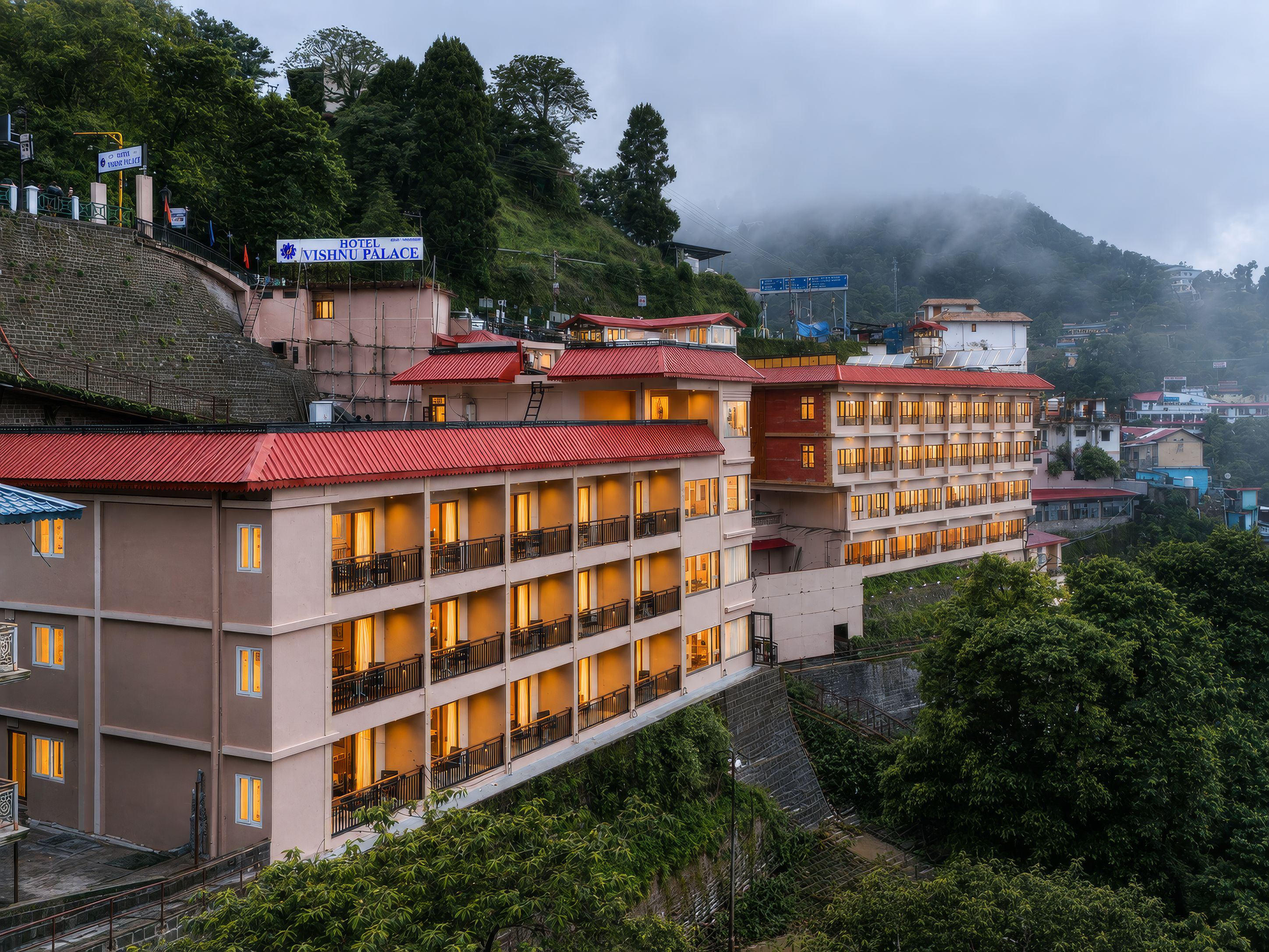 A multi-story hotel with balconies and a red roof, surrounded by lush greenery and hills.