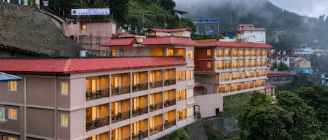 A multi-story hotel with balconies and a red roof, surrounded by lush greenery and hills.