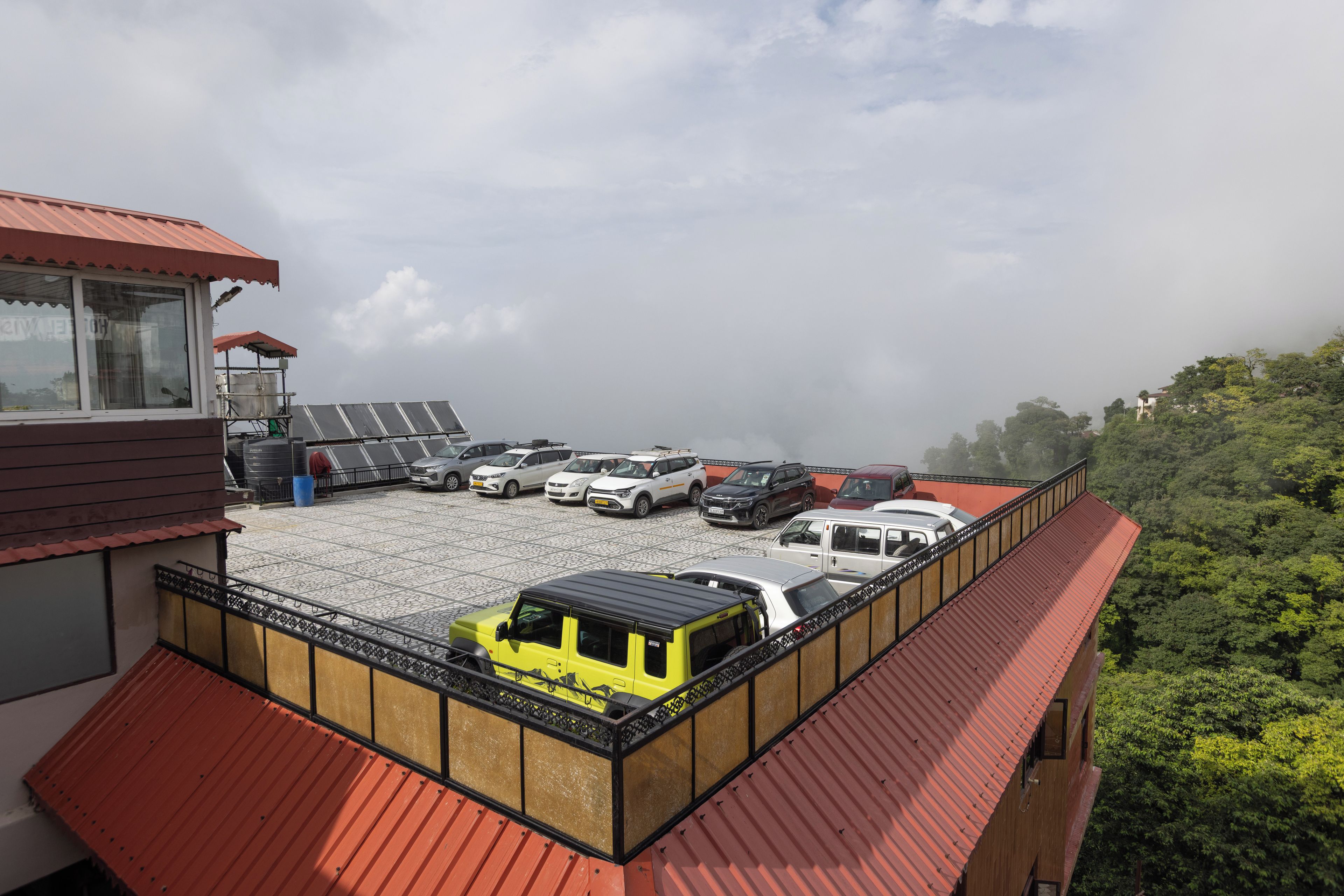 A parking area with multiple vehicles, a building with a red roof, and a view of a forested hill.