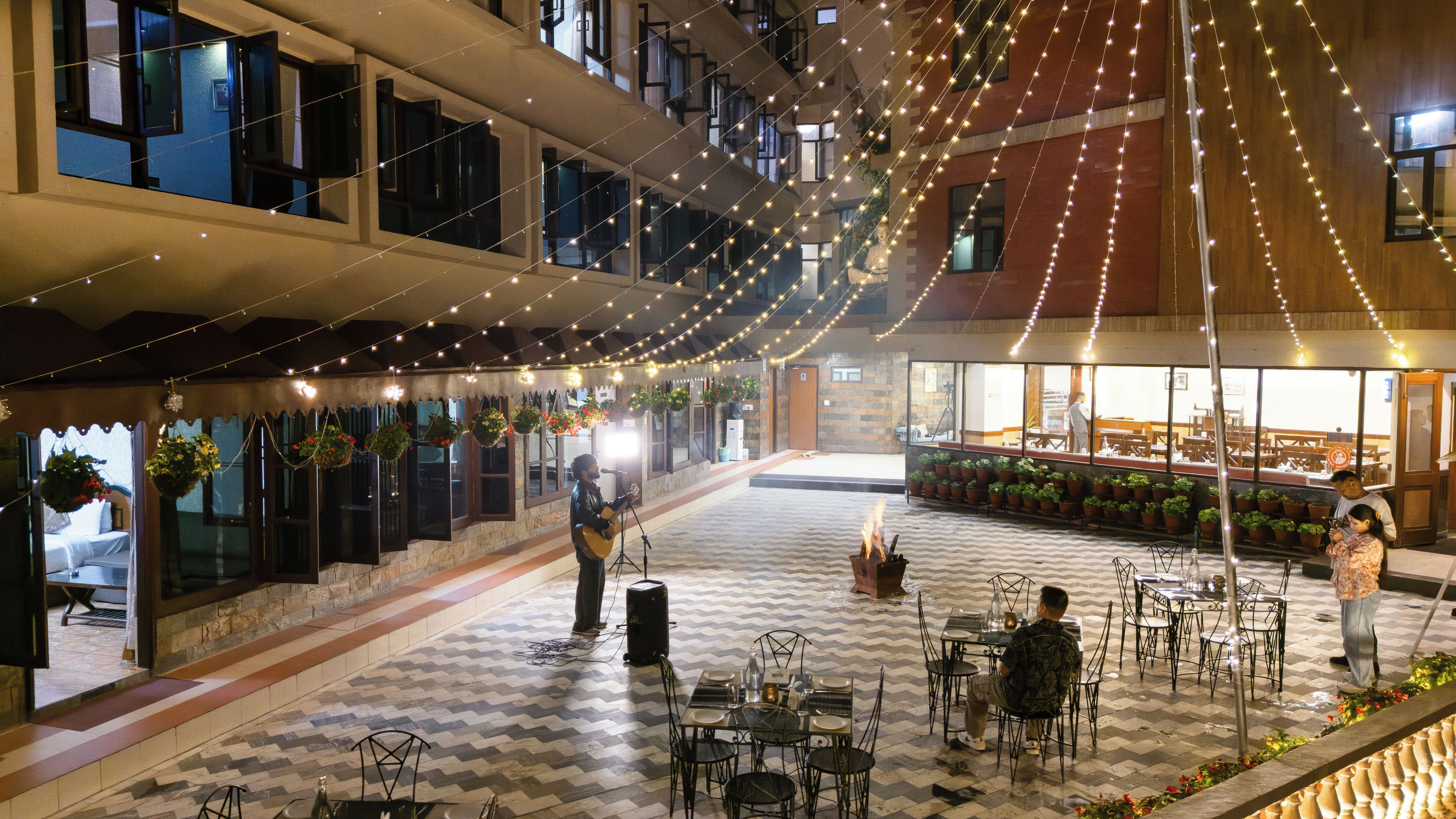 Un patio interior con luces colgantes, plantas en macetas y mesas listas para comer.