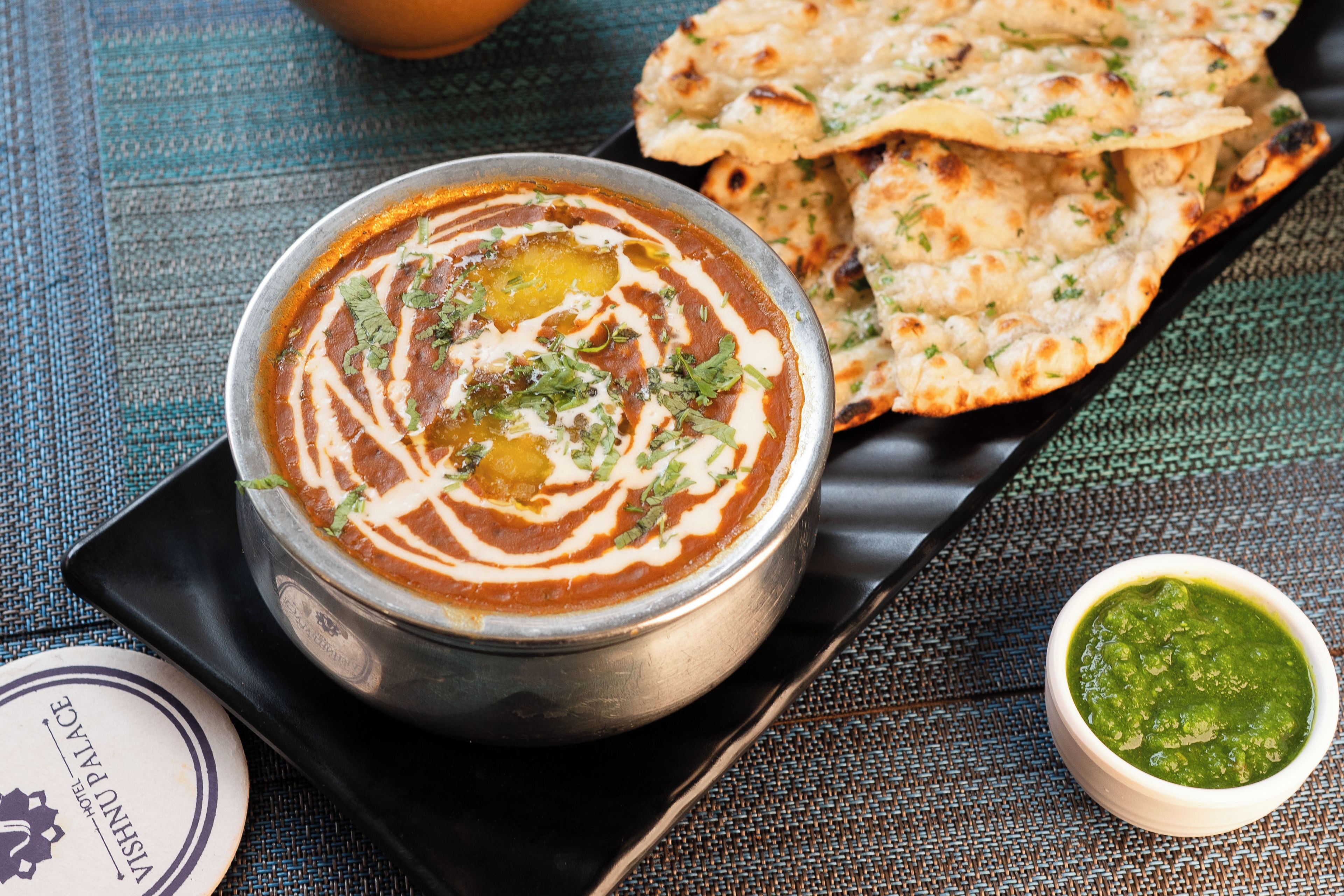 A bowl of curry with naan bread and a small bowl of chutney on a woven mat.
