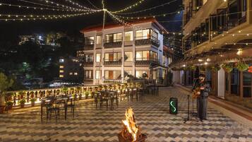 Un espacio de comedor al aire libre con mesas, sillas, un pozo de fuego y luces colgantes.