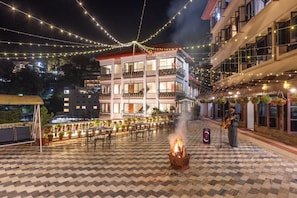 Un área de comedor al aire libre con un brasero, luces colgantes y un edificio al fondo.
