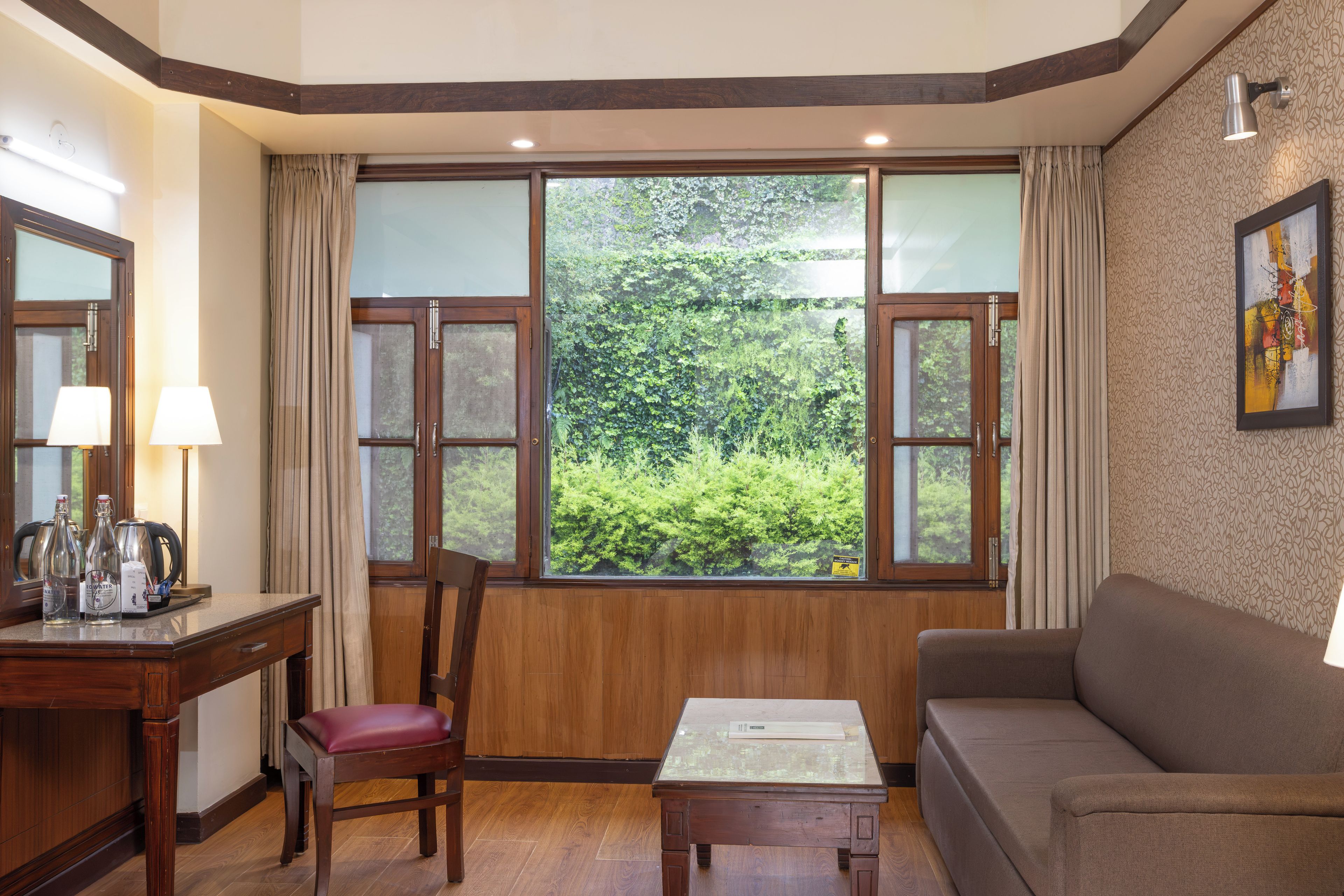 Una habitación con suelo de madera, un sofá marrón, una mesa de madera, una silla y un ventanal con vistas a zonas verdes.