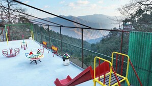 Un parque infantil con tobogán, columpios y bancos, rodeado por una valla de seguridad y con vistas a montañas al fondo.