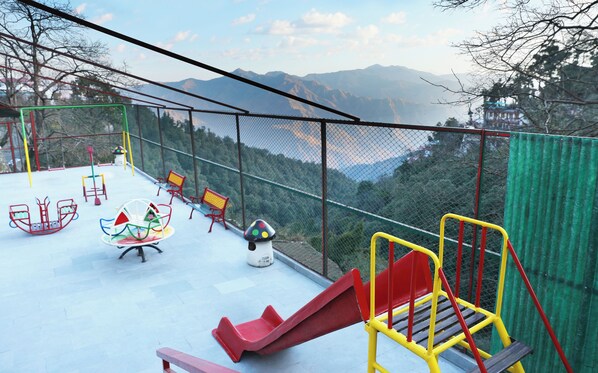 A playground with a slide, swings, and benches, surrounded by a safety fence with a view of mountains in the background.