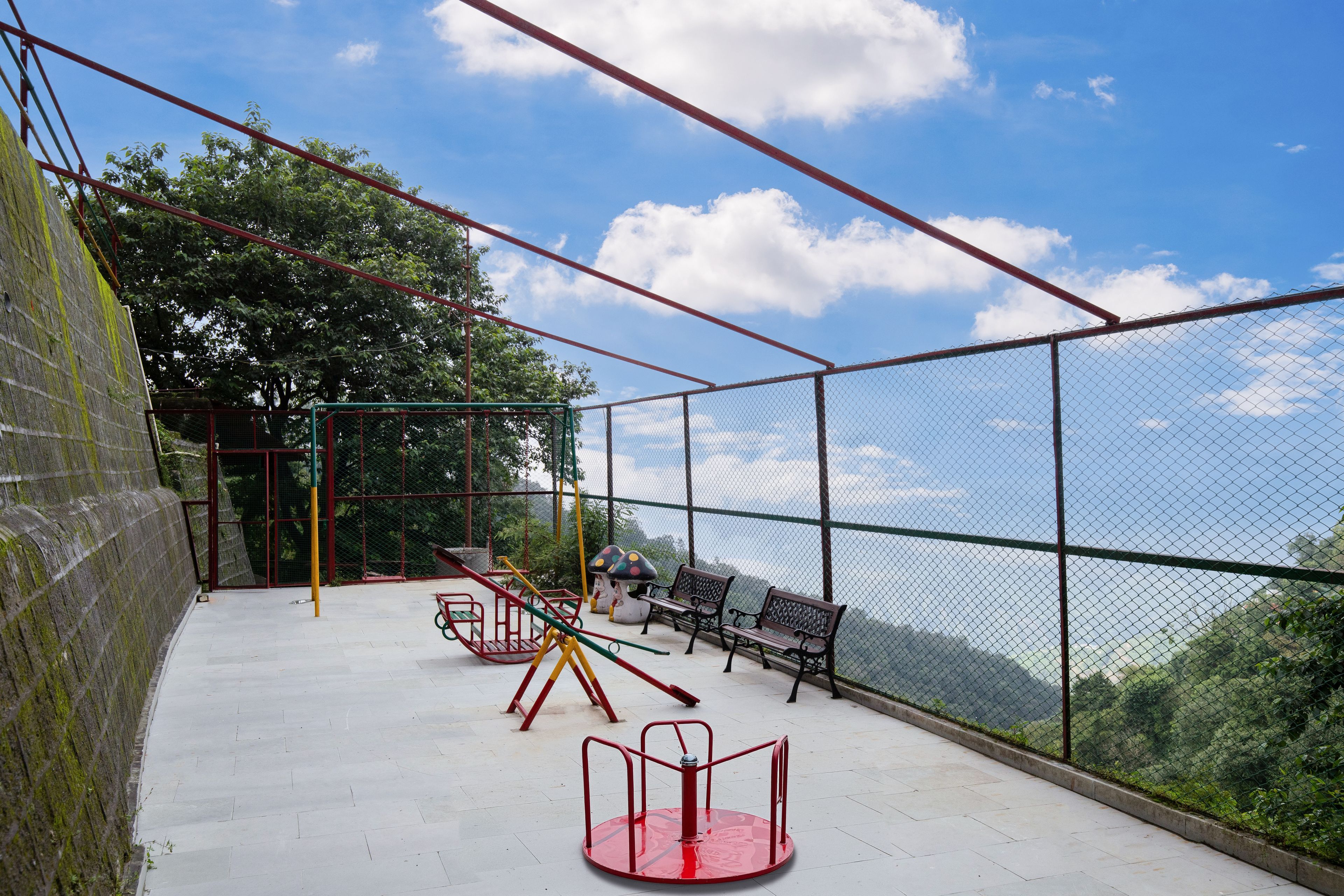 A playground with swings, a slide, and a bench, enclosed by a fence with a view of trees and sky.