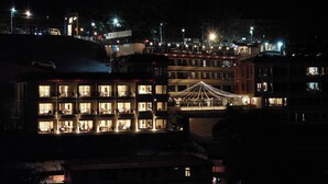 Un hotel de varios pisos con ventanas e balcones iluminados, un área de estar al aire libre cubierta y vistas a una colina con luces.