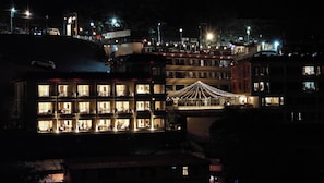 Un hôtel à plusieurs étages, doté de fenêtres et de balcons illuminés, d’un espace extérieur couvert avec des sièges, et offrant une vue sur une colline parsemée de lumières.
