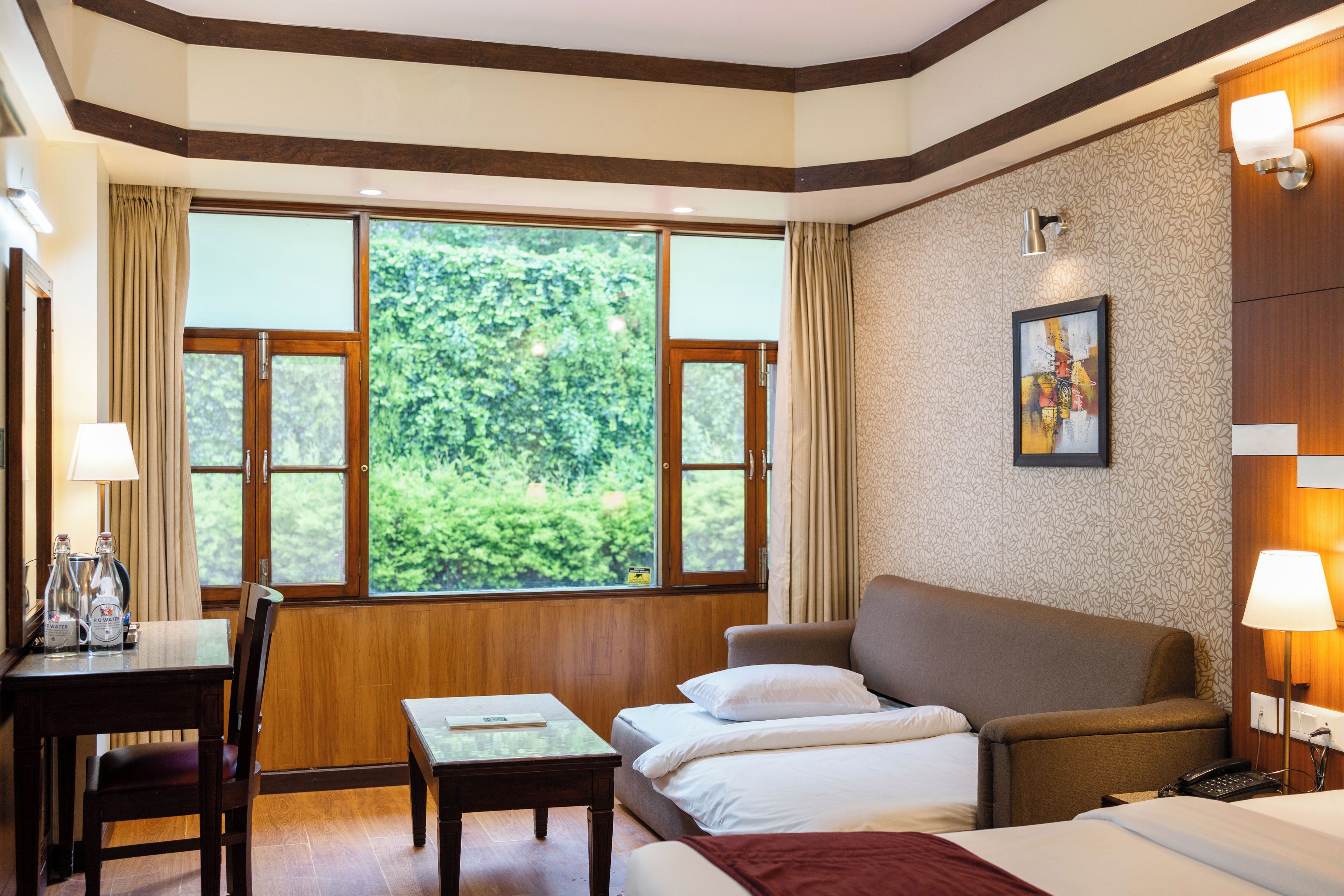 Habitación de hotel con un ventanal grande, una cama, un sofá, una mesa con lámpara y un cuadro en la pared.