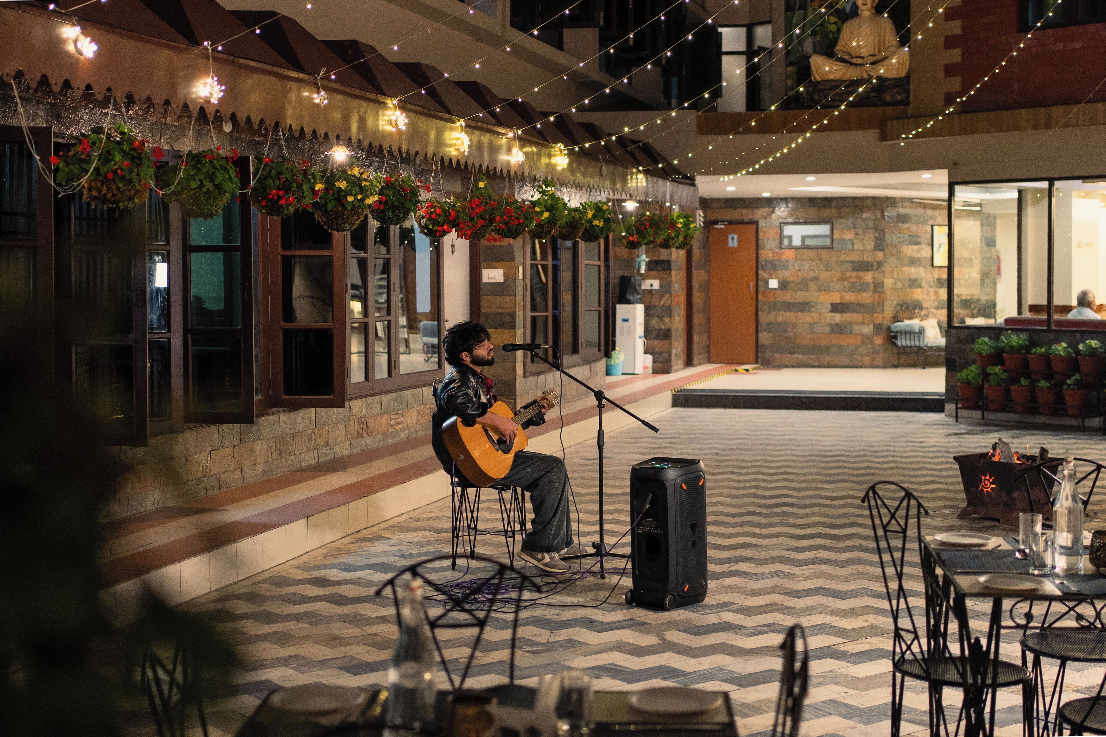 Um músico se apresentando em um terraço ao ar livre, com um suporte de microfone e um alto-falante, cercado por jardineiras suspensas e luzes de Natal.