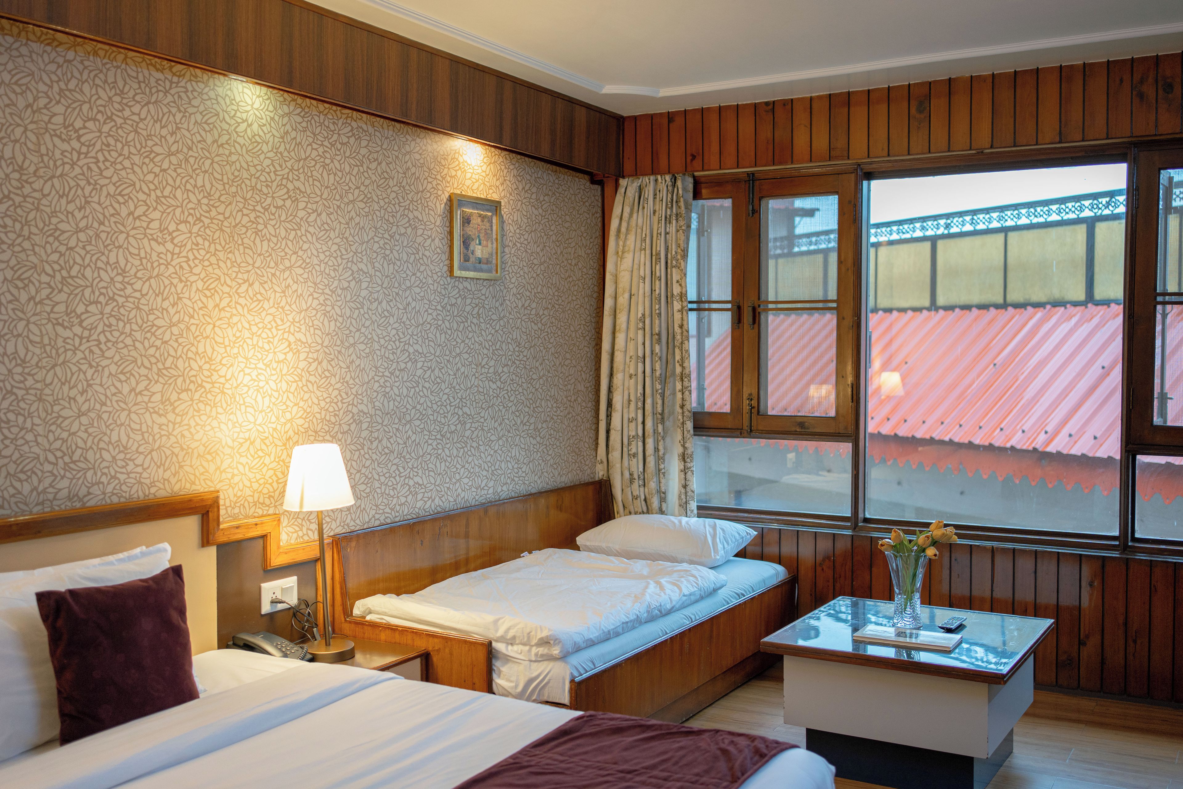 A hotel room with a bed, bedside table, lamp, and a window with a view of a red-roofed building.