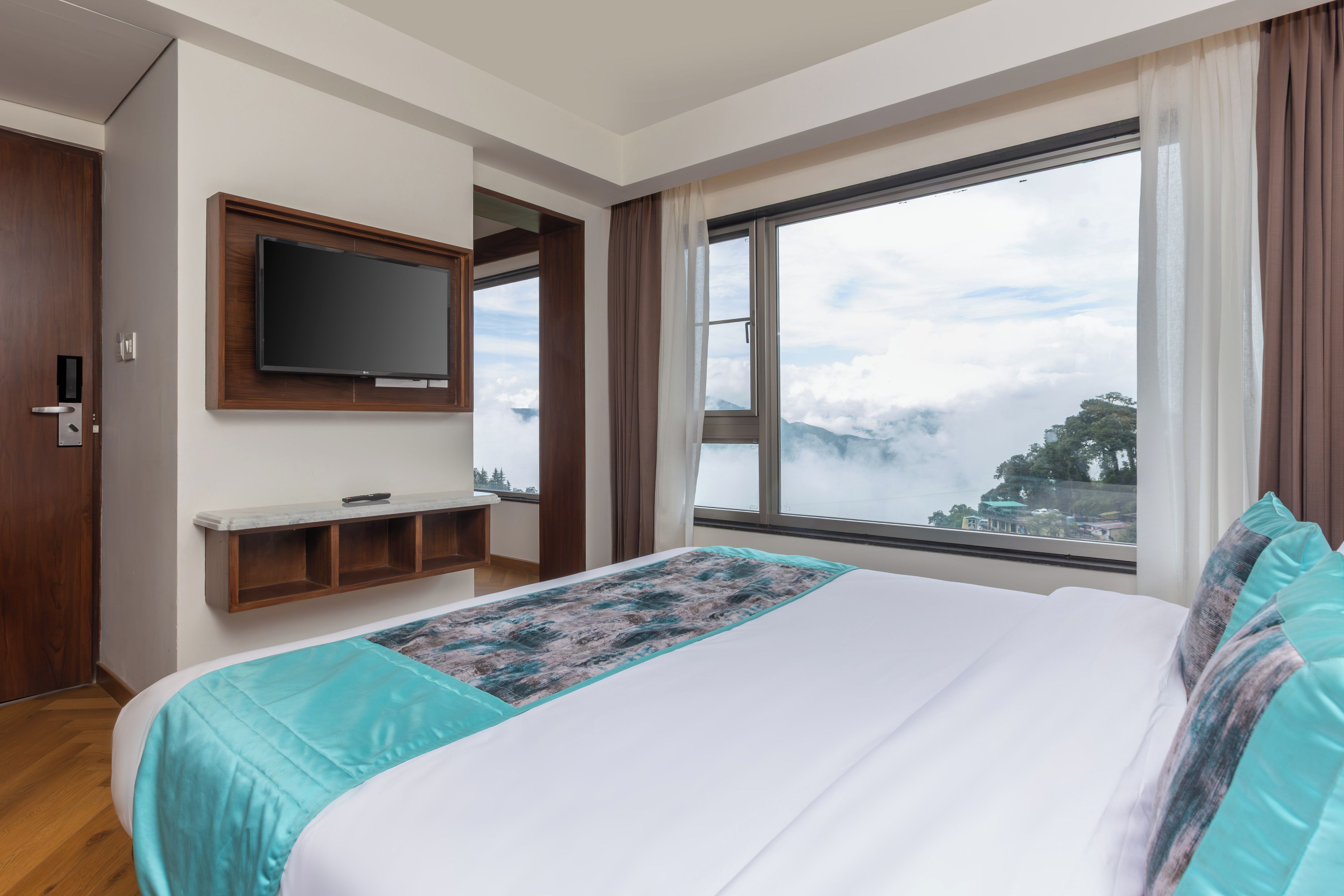 A hotel room with a bed, a television mounted on the wall, a window with a view of clouds and trees, and a wooden door.