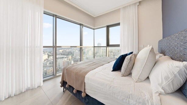 A modern bedroom with a large bed, white bedding, and a city view through large windows.