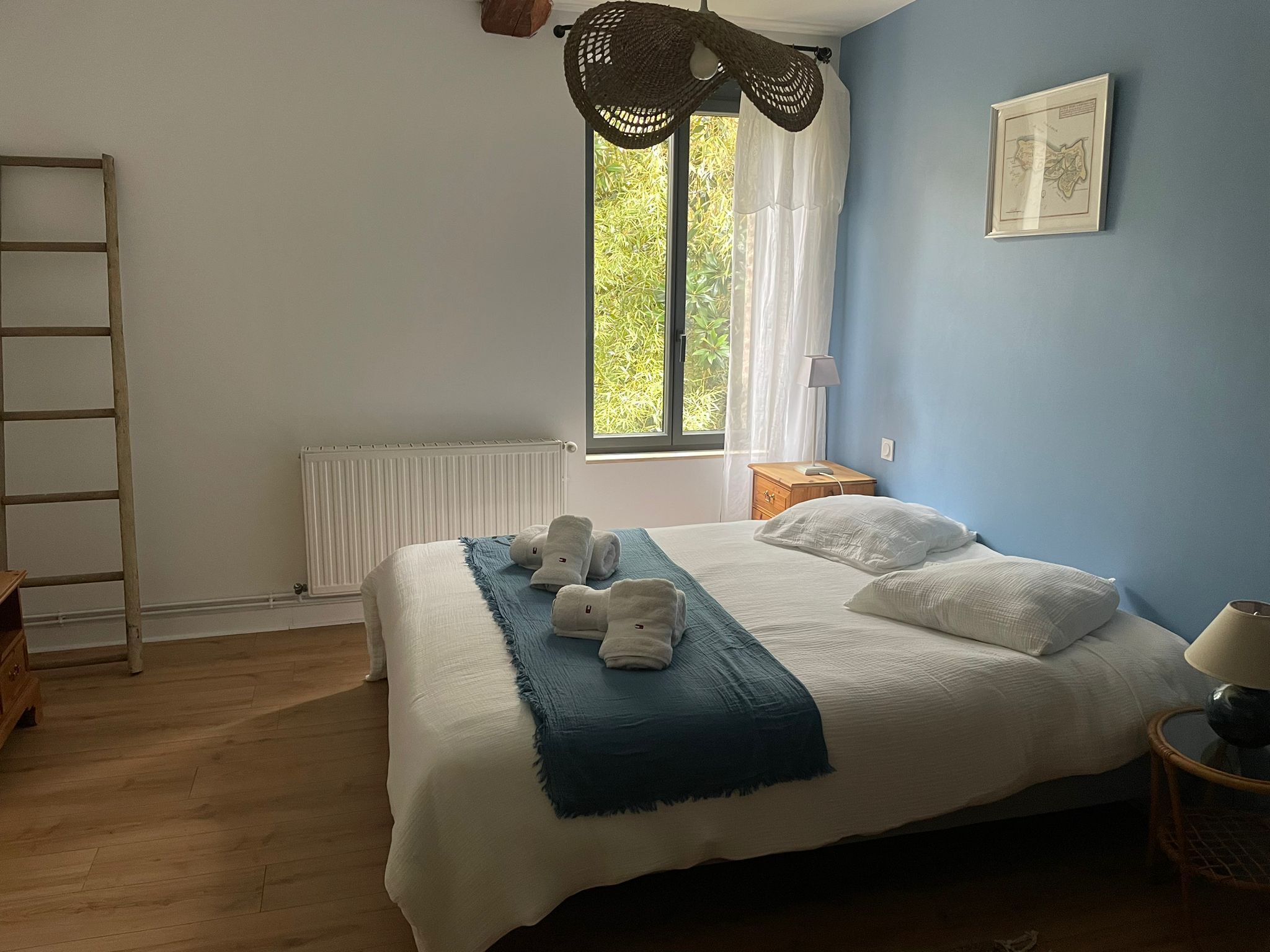 A bedroom with a bed, a window with curtains, a radiator, a wooden ladder, a bedside table with a lamp, and a framed picture on the wall.