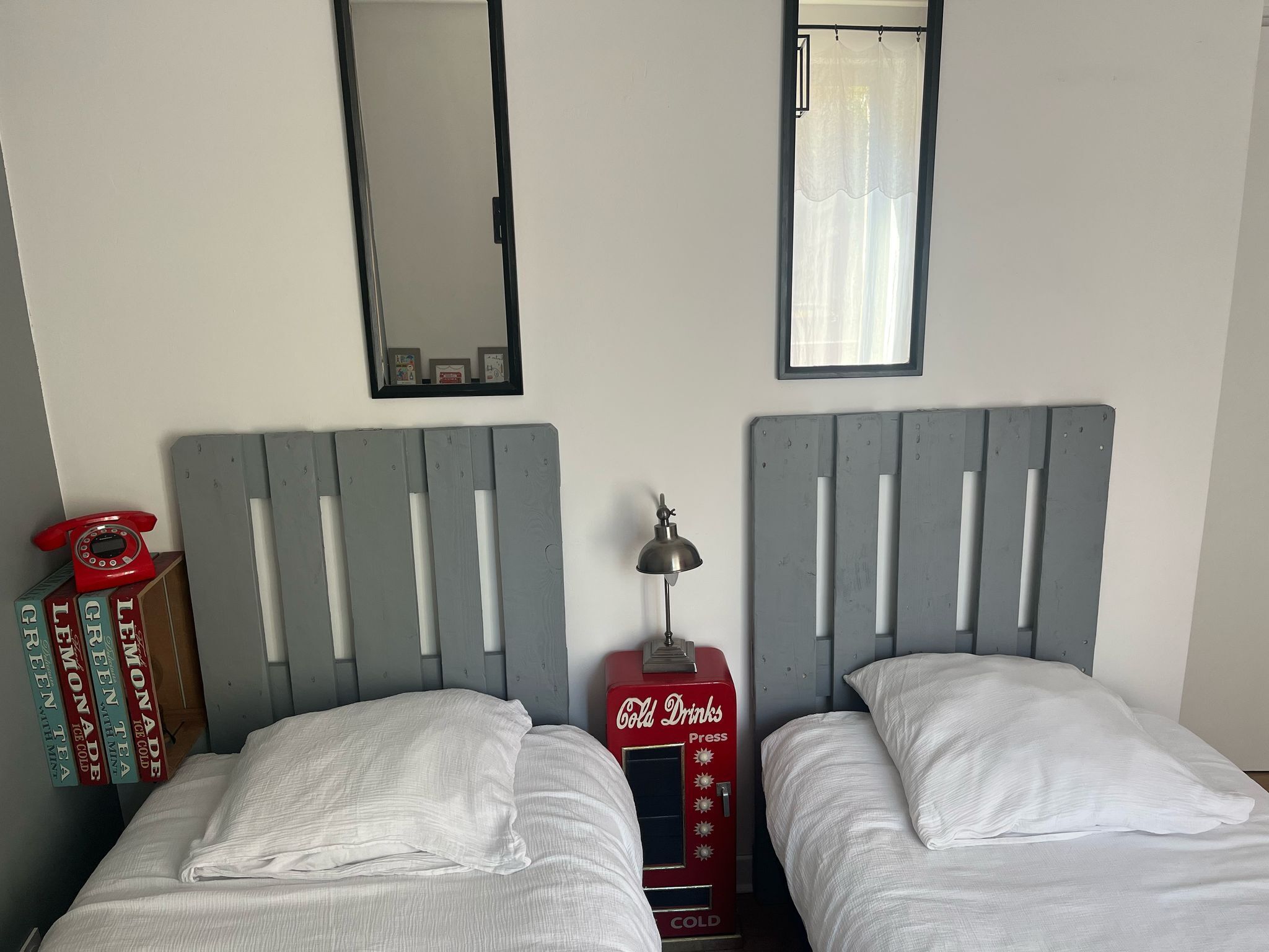 A room with two beds, a mirror, a window with a curtain, and a vintage Coca-Cola vending machine.