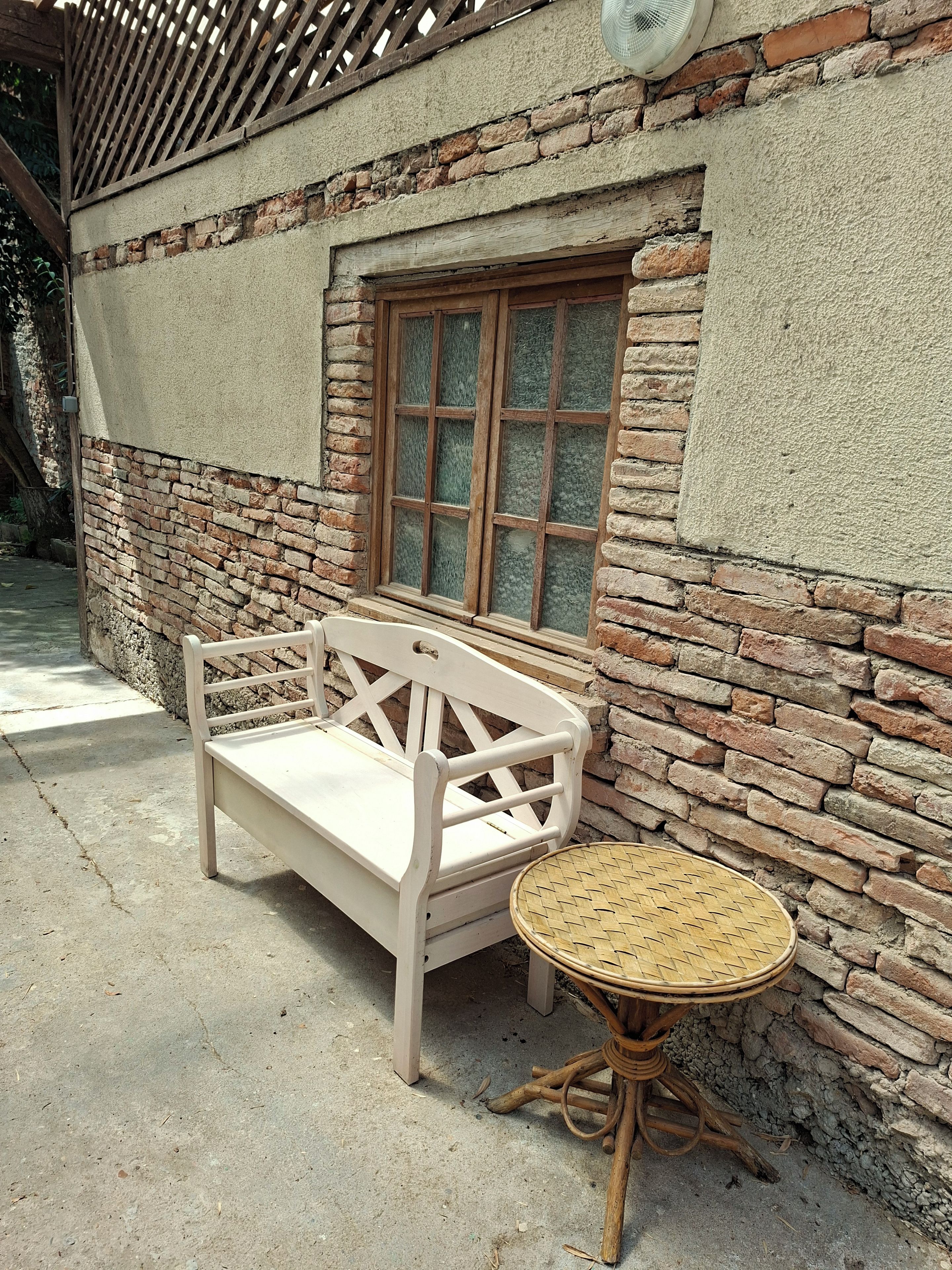 Un banc blanc et une table ronde dont le plateau est fait d’un matériau tressé, placés contre un mur de briques percé d’une fenêtre.