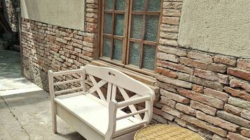 Un banc blanc et une table ronde dont le plateau est fait d’un matériau tressé, placés contre un mur de briques percé d’une fenêtre.