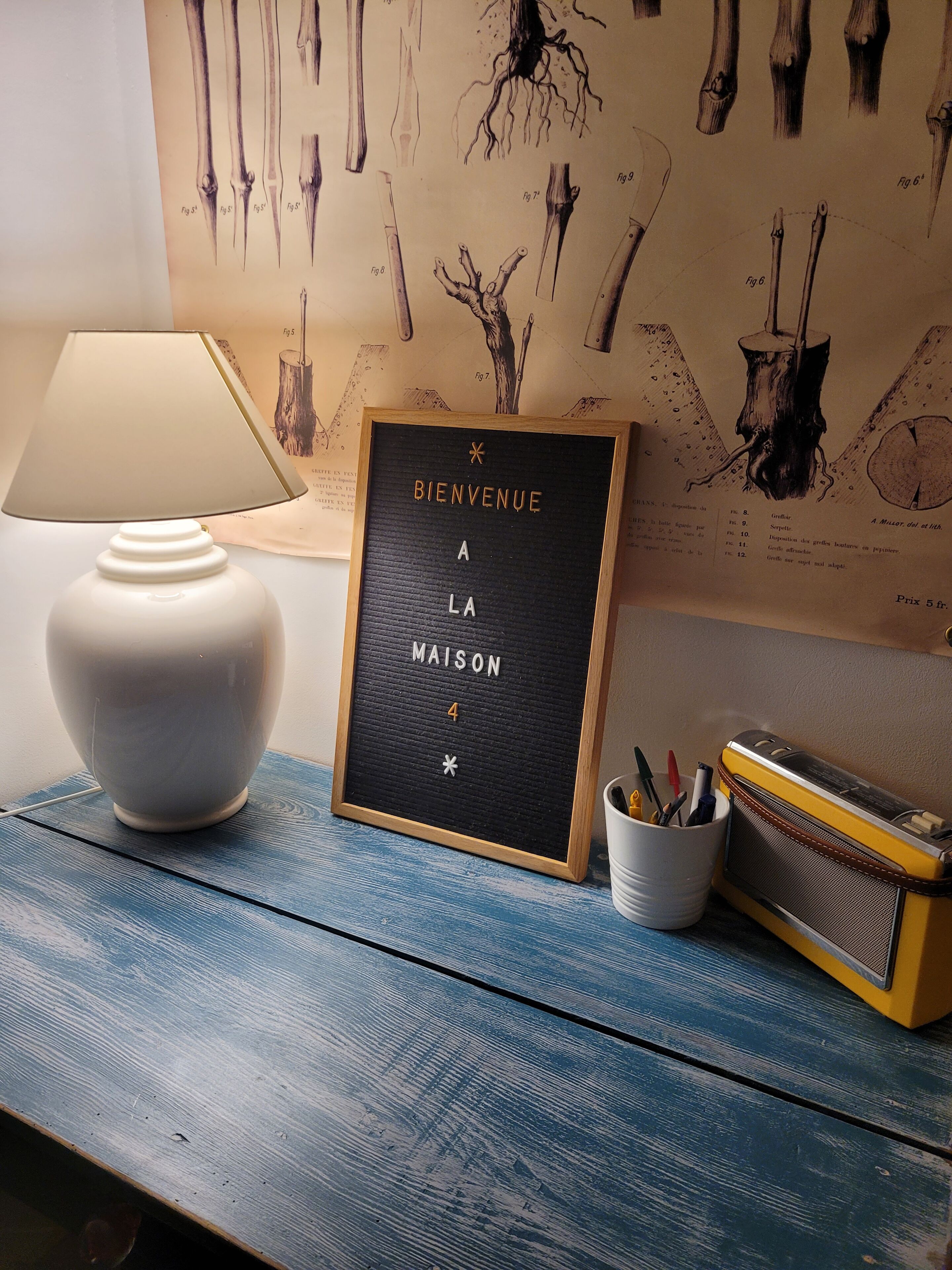 Et indrammet velkommensskilt med teksten "Bienvenue A La Maison 4" i et rum med en lampe, en vintage radio og en væg med anatomiske tegninger.