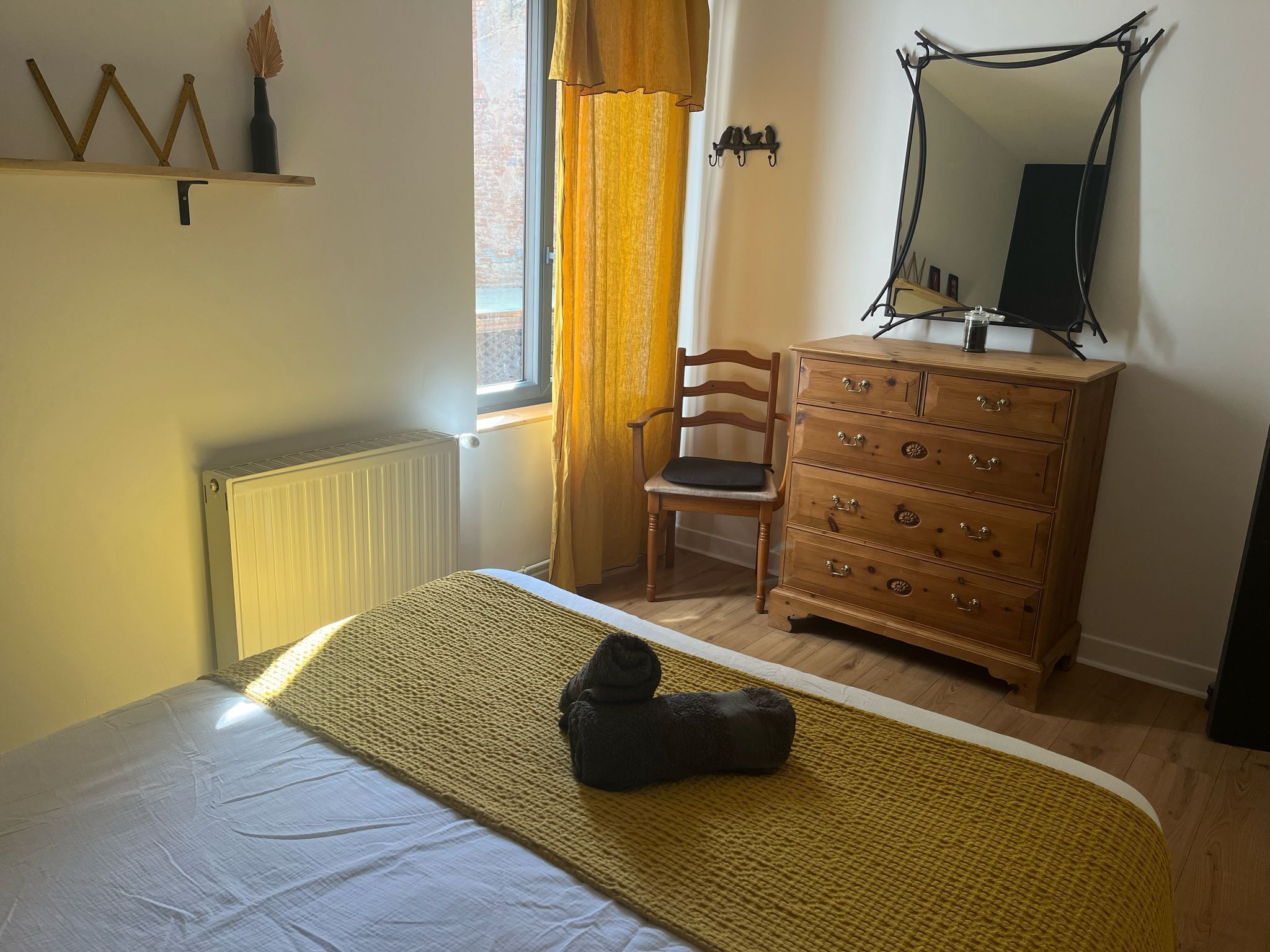 Une chambre à coucher avec un lit, une commode en bois, une chaise, un radiateur et une fenêtre avec des rideaux.