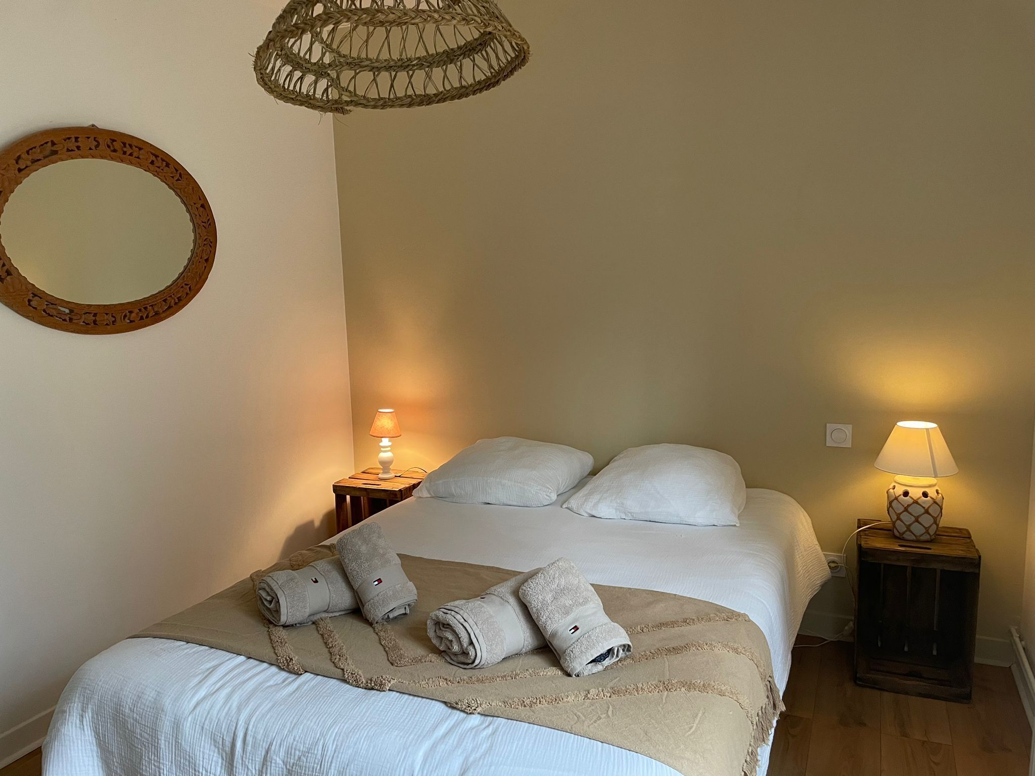 A neatly made bed with pillows and rolled towels, a round mirror, and two bedside lamps on wooden nightstands.