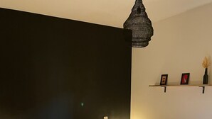 Ein ordentlich bezogenes Bett mit weißer Decke und einem senffarbenen Überwurf, eine hängende Pendelleuchte sowie ein kleiner Nachttisch mit Lampe.
