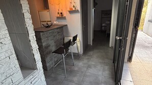 A modern interior with a stone wall, a dining area with a chair and table, and a kitchen in the background. - Maison T2 Casa Alegria Peace and greenery in the heart of the Pyrénées-Orientales  (Joch)