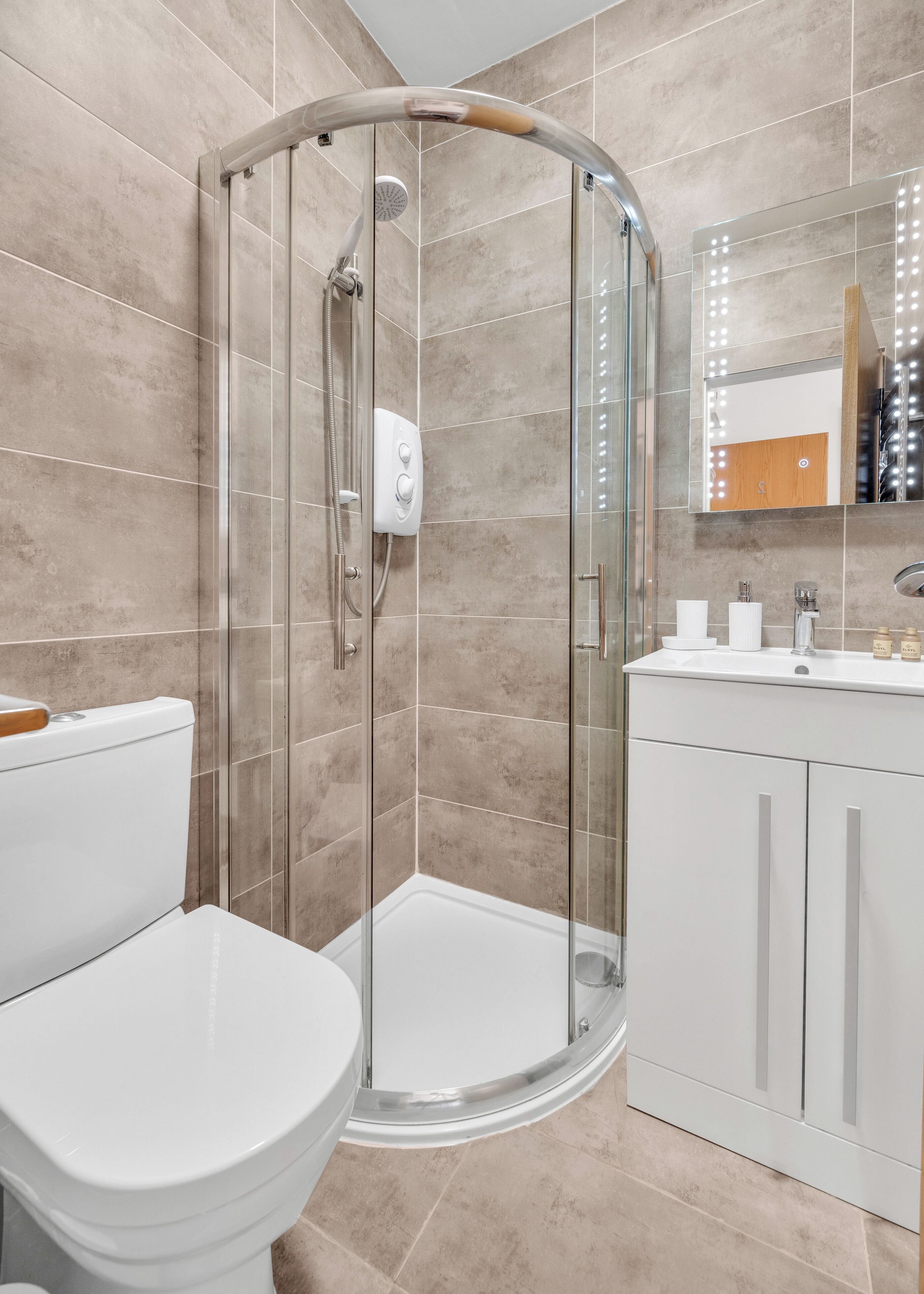 Une salle de bain moderne avec une douche à parois de verre, des toilettes blanches et un meuble-lavabo doté d’un miroir et d’un éclairage.