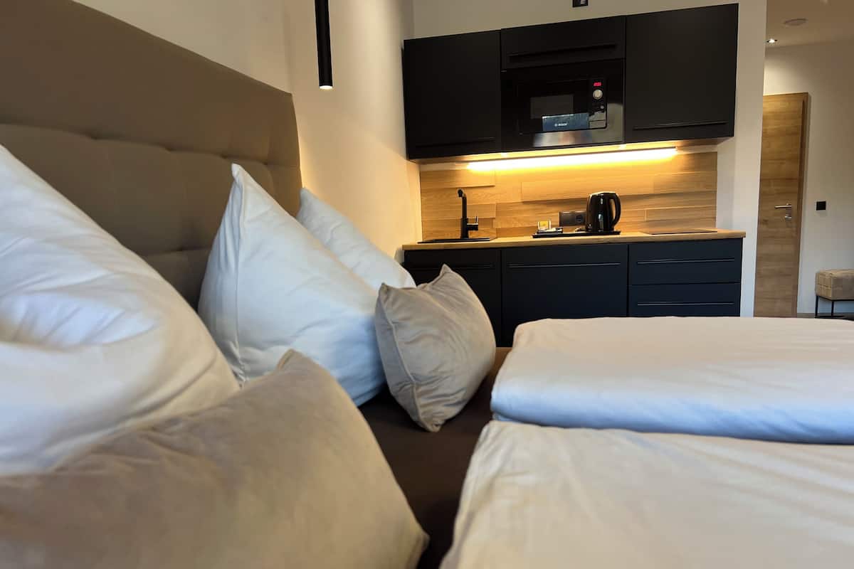 A hotel room with a bed, pillows, a bedside table, a coffee maker, and a built-in kitchen area.