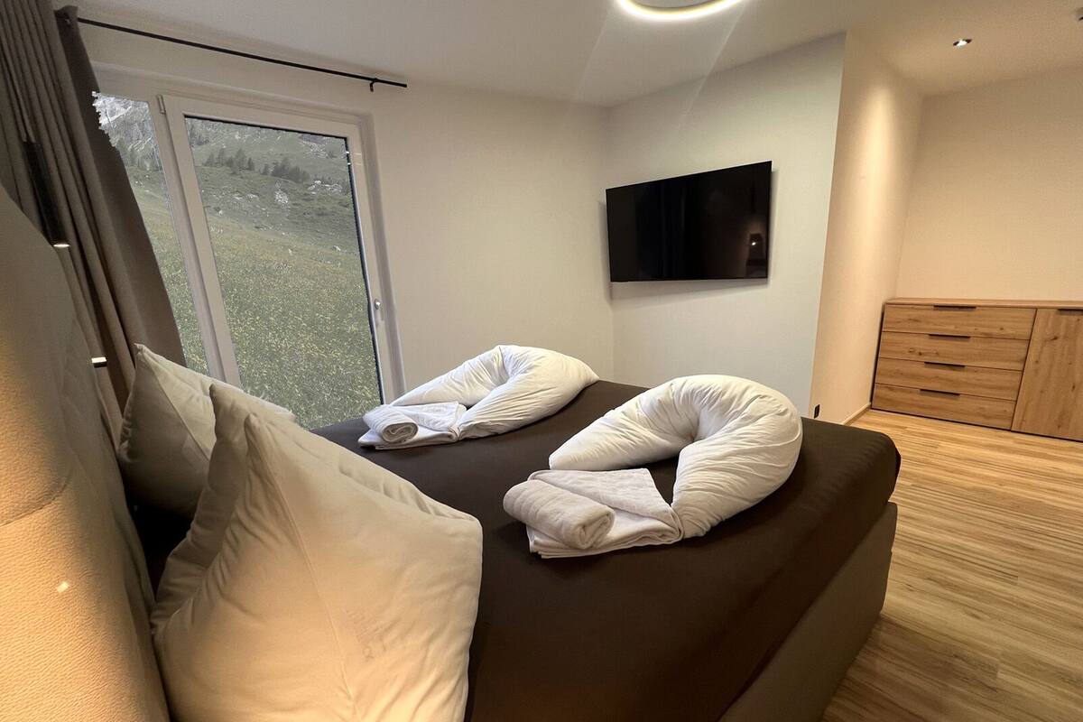 A modern room with a bed, pillows, a flat-screen TV, and a view of greenery outside.