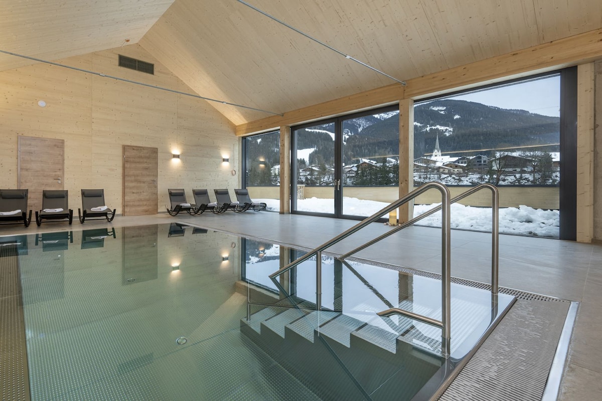 Une piscine intérieure moderne avec des murs en verre transparent, un plafond en bois et une vue sur des montagnes enneigées.
