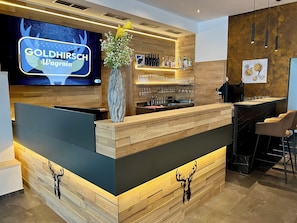 A modern bar area with a wooden reception desk, a TV screen displaying the name 'GOLDHIRSCH Wagrain,' and a vase with flowers. - Goldhirsch Aparthotel Wagrain (Wagrain)